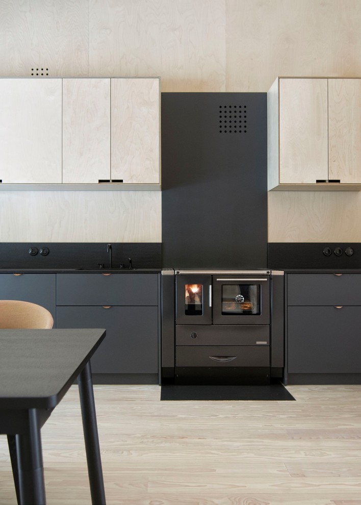 Photo of a mid-sized single-wall open plan kitchen with an undermount sink, flat-panel cabinets, light wood cabinets, quartz benchtops, black splashback, black appliances, painted wood floors, no island and beige floor.