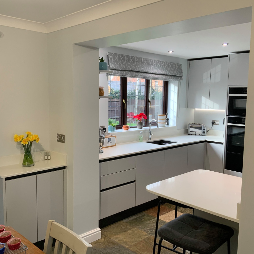 Inspiration for a mid-sized contemporary eat-in kitchen in Buckinghamshire with flat-panel cabinets, grey cabinets, quartzite benchtops, metallic splashback, mirror splashback, black appliances, a peninsula and white benchtop.