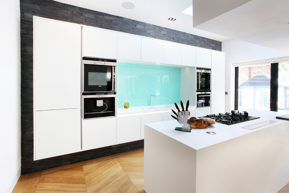Inspiration for a mid-sized contemporary galley kitchen in Manchester with flat-panel cabinets, white cabinets, light hardwood floors, with island, a double-bowl sink, blue splashback, glass sheet splashback and stainless steel appliances.