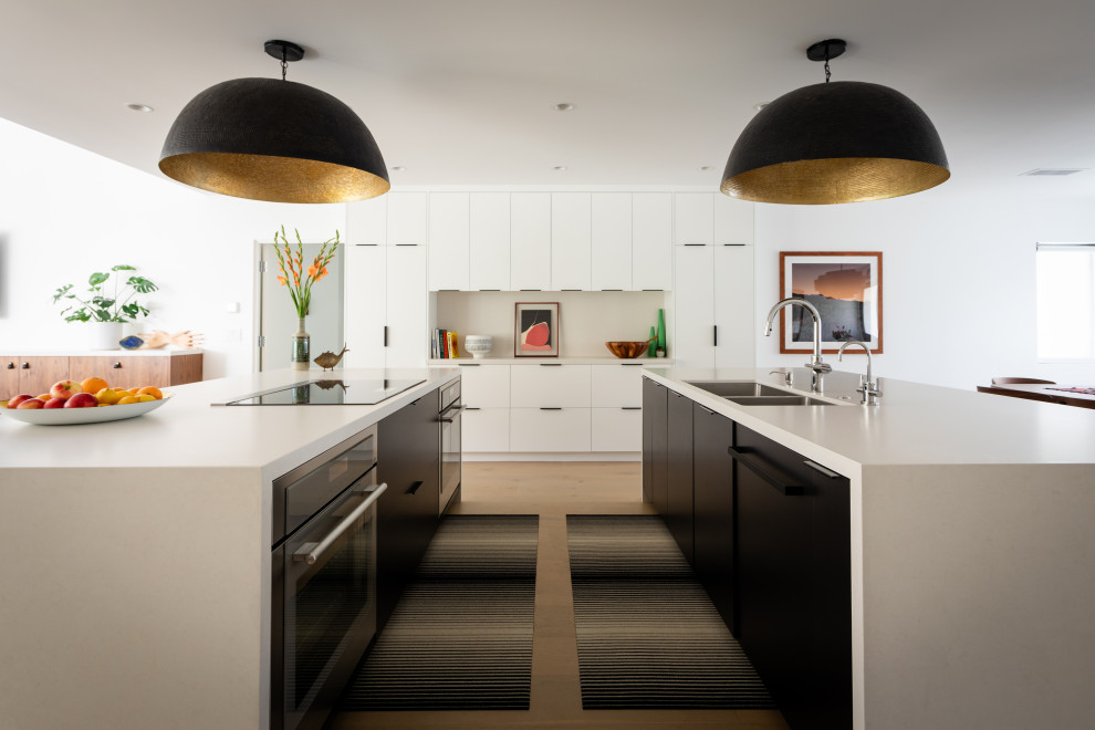 Photo of a midcentury u-shaped kitchen in Santa Barbara with multiple islands, an undermount sink, flat-panel cabinets, white cabinets, stainless steel appliances, light hardwood floors, beige floor and white benchtop.