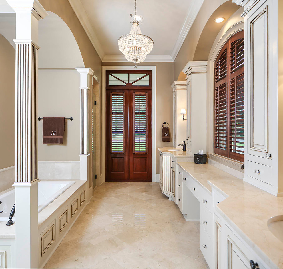 Large traditional master bathroom in New Orleans with an undermount sink, white cabinets, a drop-in tub, beige tile, beige walls, recessed-panel cabinets, marble benchtops, travertine floors and travertine.