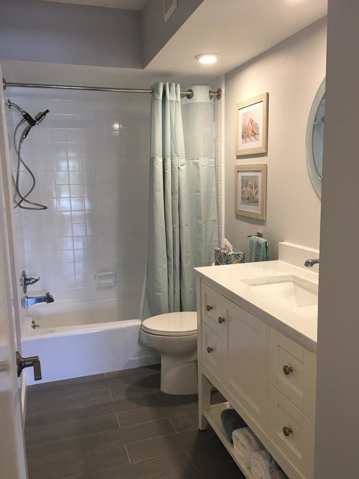 Photo of a large beach style 3/4 bathroom in Tampa with raised-panel cabinets, white cabinets, a drop-in tub, an open shower, a one-piece toilet, white tile, stone tile, grey walls, dark hardwood floors, a drop-in sink and solid surface benchtops.