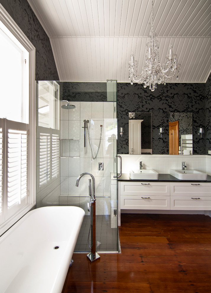 Photo of a mid-sized transitional master bathroom in Wellington with shaker cabinets, white cabinets, a claw-foot tub, a corner shower, a wall-mount toilet, black walls, medium hardwood floors, a vessel sink, engineered quartz benchtops, brown floor and a hinged shower door.