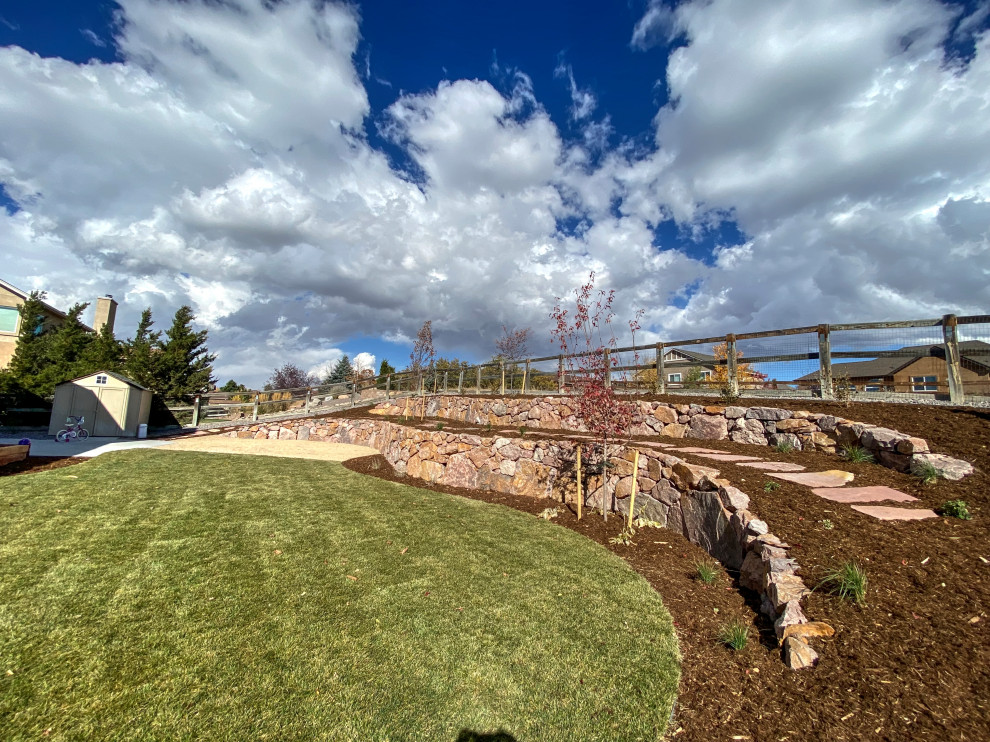 This is an example of a large rustic sloped xeriscape full sun garden for summer in Denver with a retaining wall, concrete paving and a wood fence.