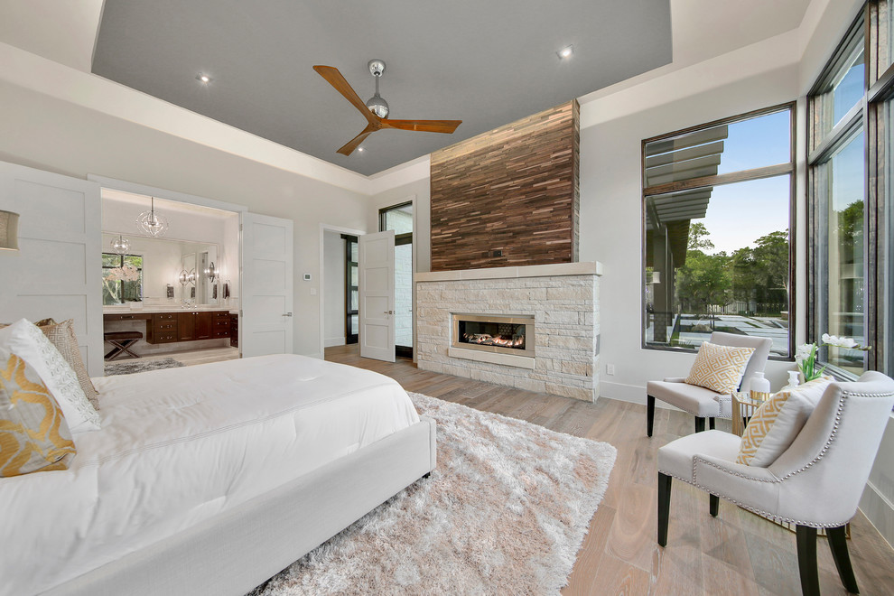 Photo of a large transitional master bedroom in Austin with grey walls, light hardwood floors, a ribbon fireplace, a stone fireplace surround and brown floor.