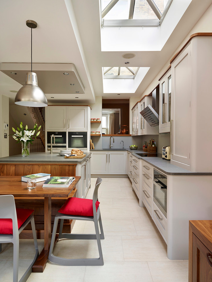 Mid-sized transitional l-shaped open plan kitchen in London with shaker cabinets, stainless steel appliances, with island, a drop-in sink, grey cabinets, grey splashback, glass sheet splashback and limestone floors.