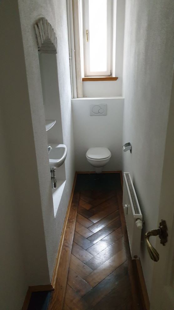 Kleine Urige Gästetoilette mit Wandtoilette mit Spülkasten, weißer Wandfarbe, braunem Holzboden und Wandwaschbecken in München