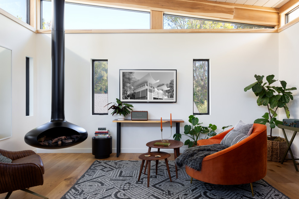 Example of a large 1950s light wood floor, brown floor and exposed beam living room design in Los Angeles with white walls, a hanging fireplace and a wall-mounted tv