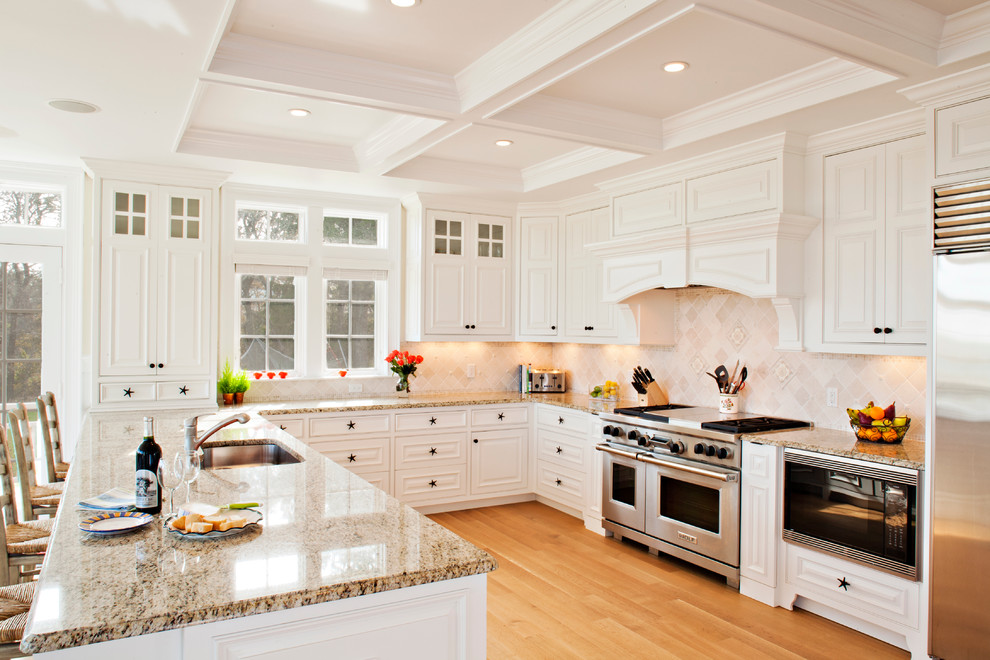 Design ideas for a beach style u-shaped eat-in kitchen in Boston with an undermount sink, recessed-panel cabinets, white cabinets, granite benchtops, beige splashback, ceramic splashback, stainless steel appliances and light hardwood floors.