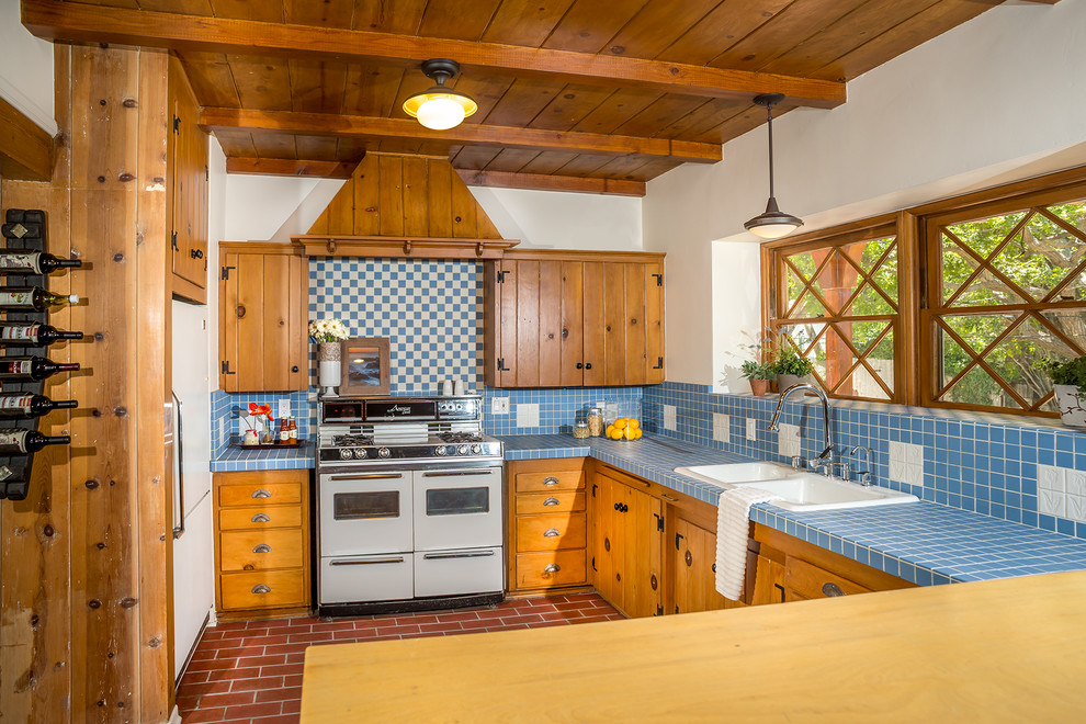Inspiration for a mid-sized traditional l-shaped kitchen in Los Angeles with a drop-in sink, medium wood cabinets, tile benchtops, white appliances, shaker cabinets, blue splashback, porcelain splashback, brick floors and blue benchtop.