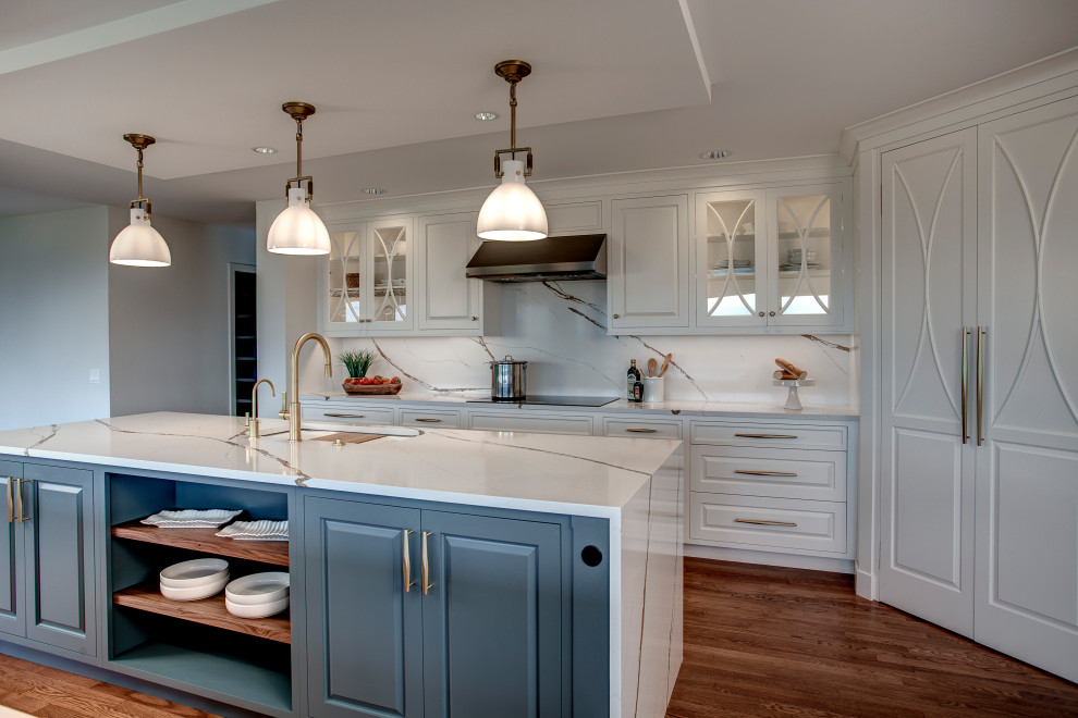 Photo of a large transitional l-shaped open plan kitchen in Seattle with a single-bowl sink, raised-panel cabinets, white cabinets, quartz benchtops, white splashback, engineered quartz splashback, stainless steel appliances, medium hardwood floors, multiple islands, brown floor, white benchtop and recessed.