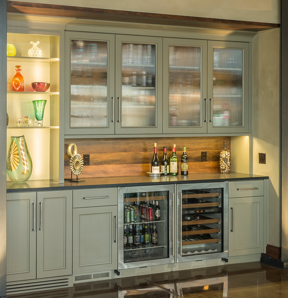 This is an example of a large traditional home bar in Chicago with grey cabinets, granite benchtops, brown splashback, ceramic splashback, concrete floors, brown floor and glass-front cabinets.