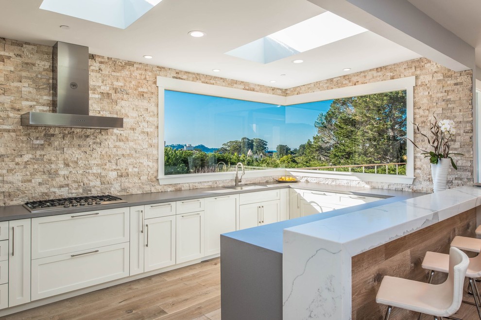 Design ideas for a large transitional u-shaped open plan kitchen in Orange County with an undermount sink, shaker cabinets, white cabinets, stone tile splashback, light hardwood floors, beige splashback, stainless steel appliances, with island and grey benchtop.