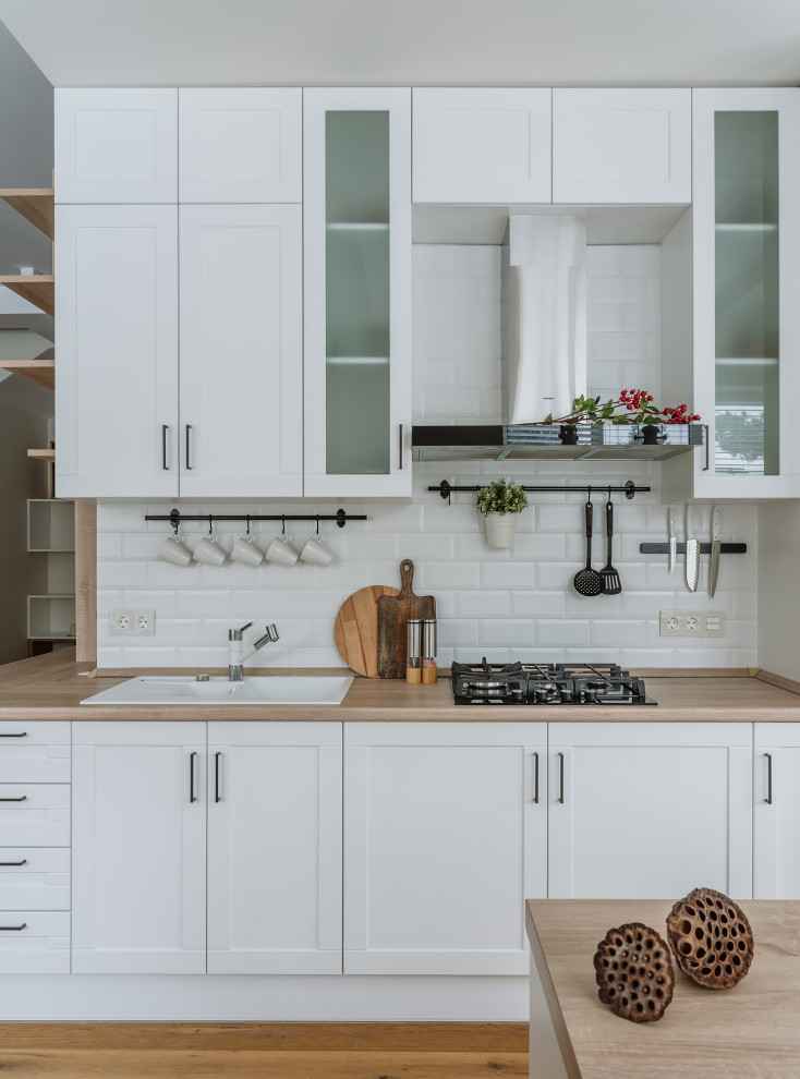 Scandinavian single-wall separate kitchen in Moscow with with island, a drop-in sink, recessed-panel cabinets, white cabinets, white splashback, subway tile splashback, medium hardwood floors, brown floor and brown benchtop.