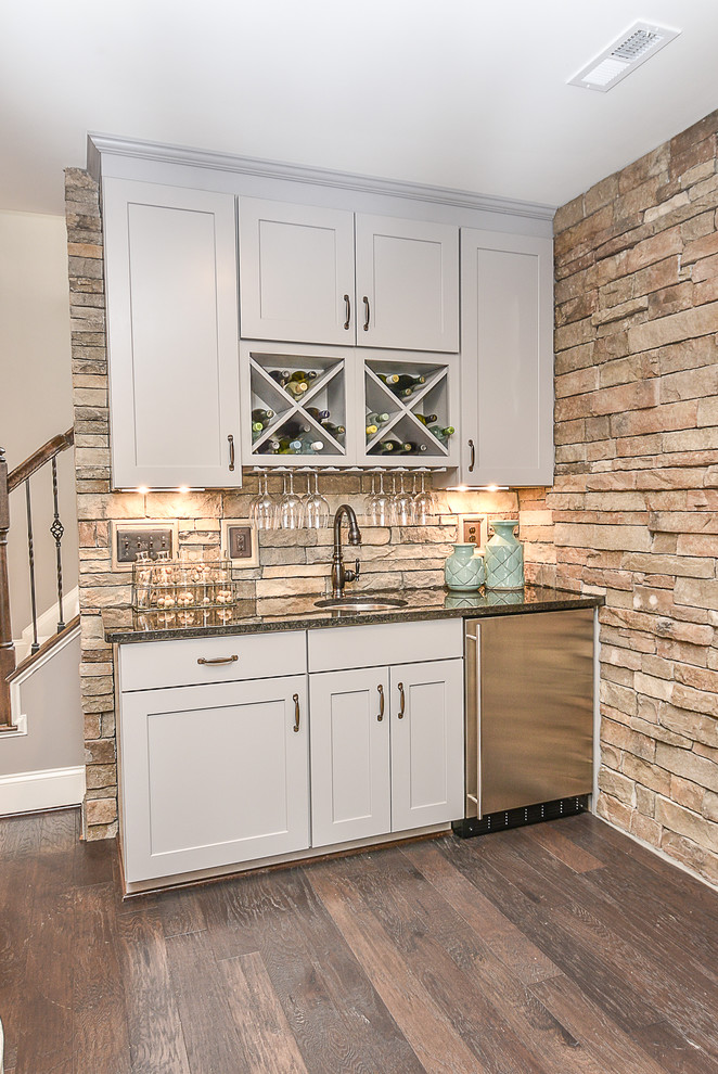 This is an example of a small transitional single-wall wet bar in DC Metro with an undermount sink, shaker cabinets, white cabinets, granite benchtops, beige splashback, stone tile splashback and dark hardwood floors.