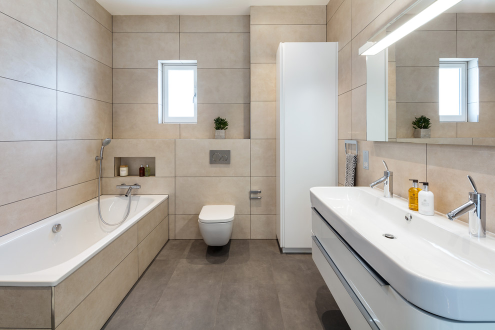 Inspiration for a mid-sized modern bathroom in London with flat-panel cabinets, white cabinets, a corner tub, a wall-mount toilet, beige tile, beige walls, a wall-mount sink and brown floor.