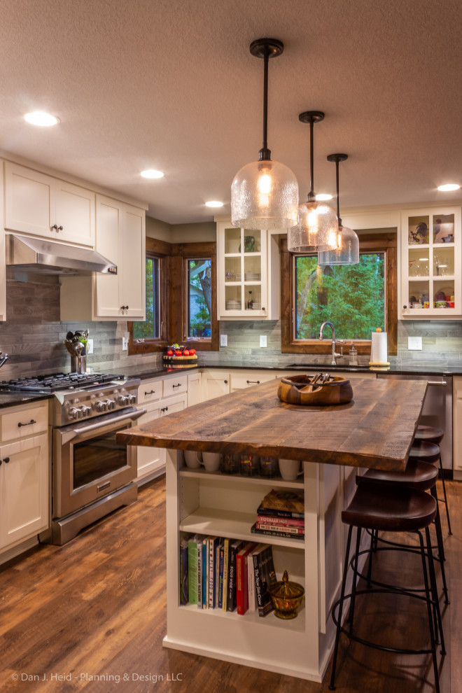 This is an example of a mid-sized country l-shaped eat-in kitchen in Minneapolis with an undermount sink, flat-panel cabinets, white cabinets, granite benchtops, grey splashback, ceramic splashback, stainless steel appliances, medium hardwood floors, with island, brown floor and black benchtop.