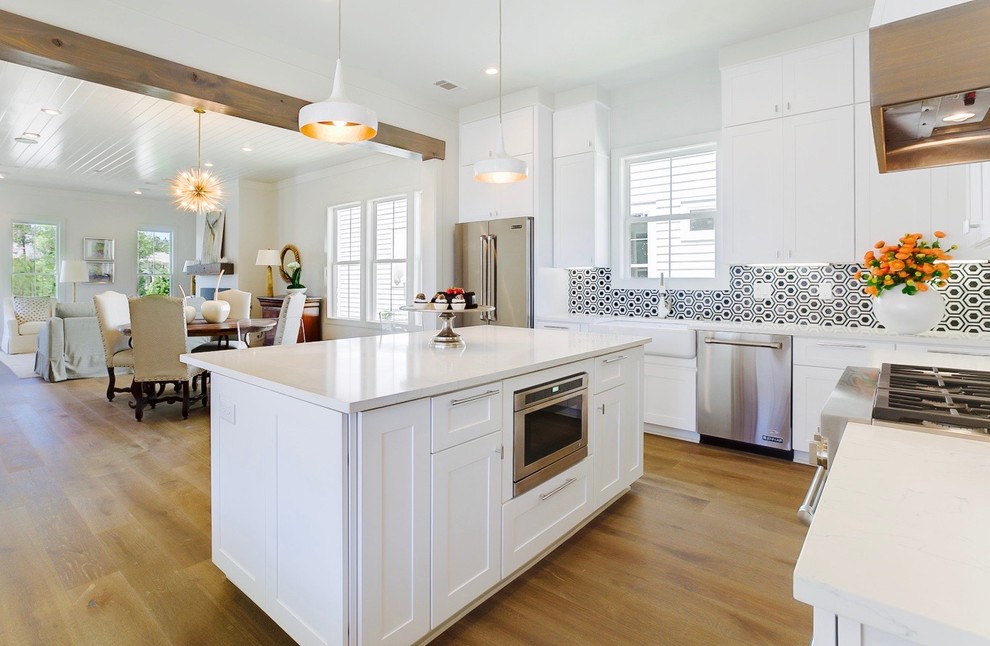This is an example of a transitional l-shaped open plan kitchen in New Orleans with a farmhouse sink, shaker cabinets, white cabinets, black splashback, stainless steel appliances, light hardwood floors and with island.