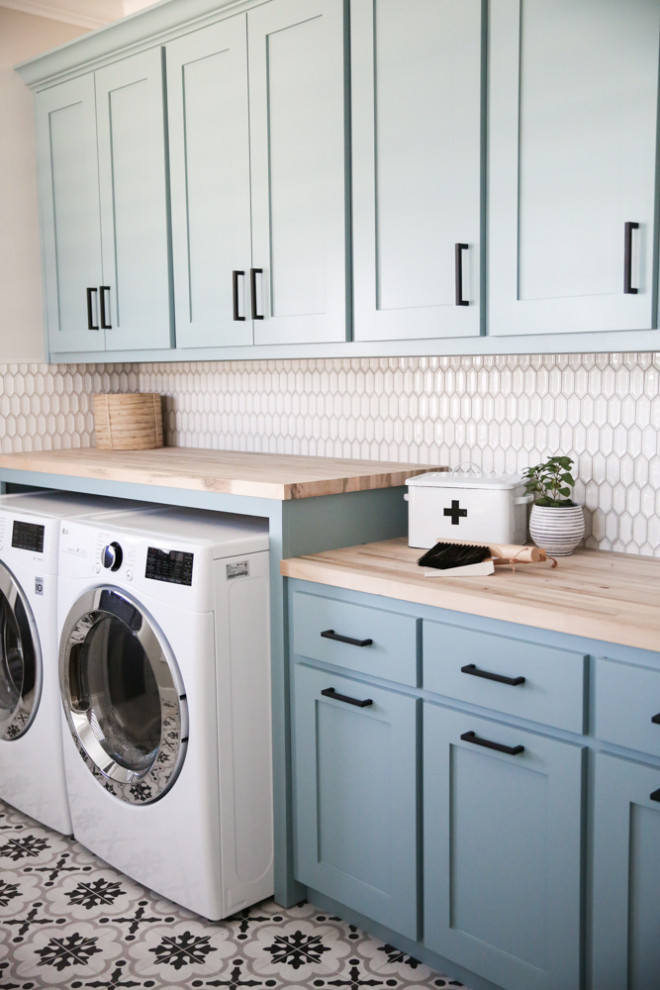 This is an example of a mid-sized traditional single-wall dedicated laundry room in Dallas with recessed-panel cabinets, blue cabinets, wood benchtops, grey walls, ceramic floors, a side-by-side washer and dryer, multi-coloured floor and brown benchtop.