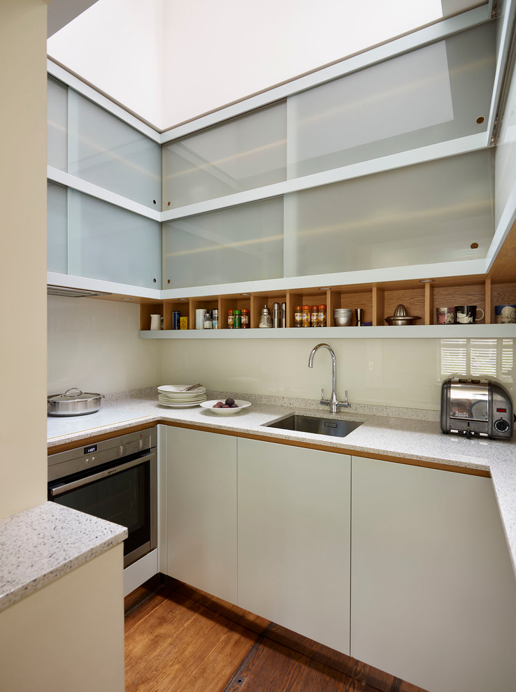 Photo of a small contemporary eat-in kitchen in London with a single-bowl sink, flat-panel cabinets, grey cabinets, solid surface benchtops, white splashback, glass sheet splashback, stainless steel appliances, medium hardwood floors and no island.