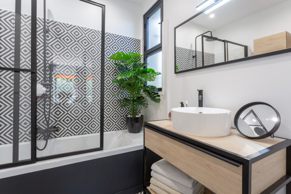 This is an example of a small contemporary master bathroom in Nice with light wood cabinets, an undermount tub, a shower/bathtub combo, a two-piece toilet, black and white tile, cement tile, white walls, cement tiles, a console sink, wood benchtops, a single vanity, a floating vanity and a sliding shower screen.