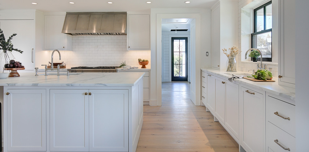 This is an example of a large traditional galley kitchen pantry in New York with a submerged sink, recessed-panel cabinets, white cabinets, engineered stone countertops, white splashback, stone tiled splashback, integrated appliances, medium hardwood flooring, an island, brown floors and white worktops.