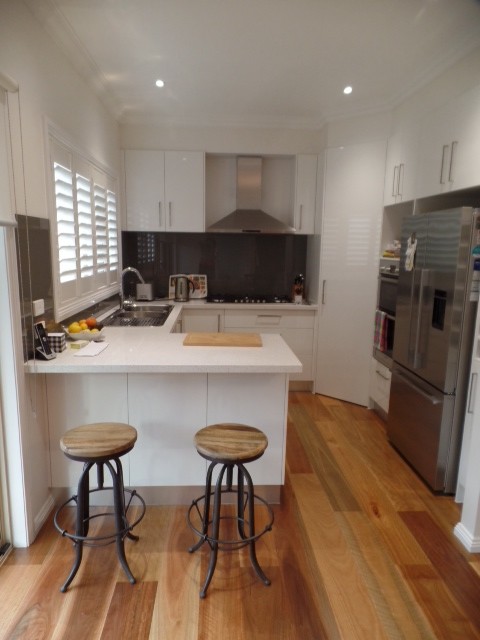 Transitional u-shaped kitchen in Melbourne with a double-bowl sink, flat-panel cabinets, white cabinets, quartz benchtops, grey splashback, glass sheet splashback, stainless steel appliances and medium hardwood floors.