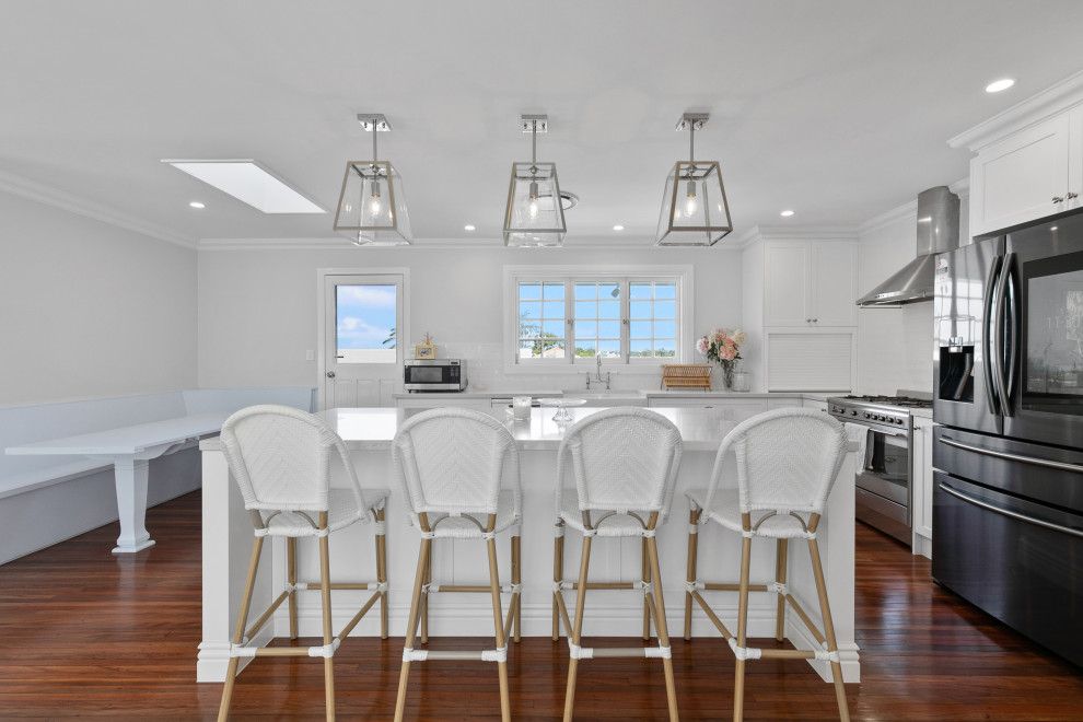 Photo of a beach style kitchen in Brisbane with a farmhouse sink, shaker cabinets, white cabinets, white splashback, stainless steel appliances, medium hardwood floors, with island and grey benchtop.