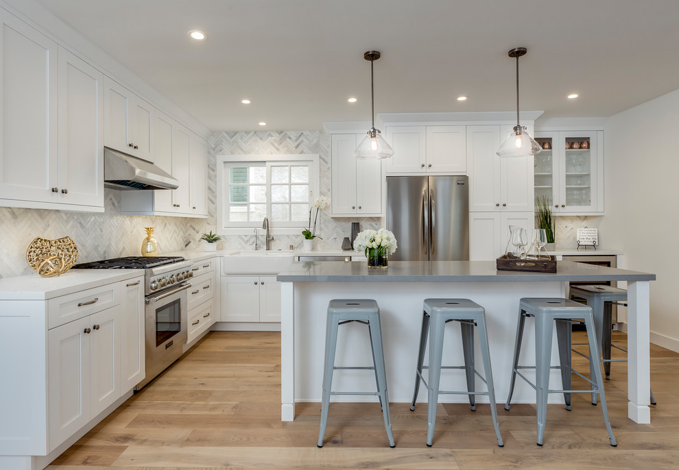 Design ideas for a large transitional l-shaped eat-in kitchen in Los Angeles with a farmhouse sink, white cabinets, white splashback, stainless steel appliances, light hardwood floors, with island, beige floor, recessed-panel cabinets, quartz benchtops and stone tile splashback.
