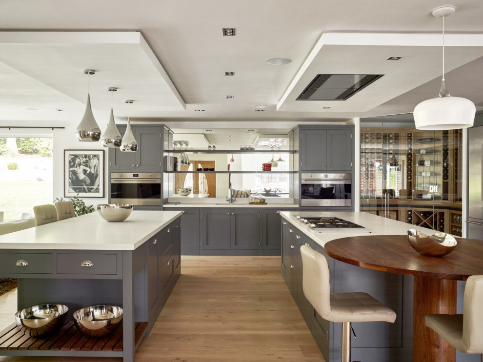 This is an example of an expansive industrial l-shaped open plan kitchen in Sussex with an undermount sink, shaker cabinets, grey cabinets, quartzite benchtops, glass sheet splashback, stainless steel appliances, light hardwood floors, multiple islands, white benchtop and coffered.