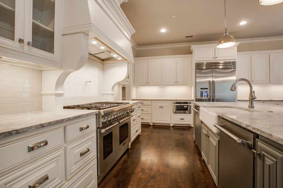 This is an example of a large traditional l-shaped eat-in kitchen in Dallas with raised-panel cabinets, white cabinets, marble benchtops, white splashback, stainless steel appliances, medium hardwood floors, with island, a farmhouse sink, subway tile splashback and brown floor.