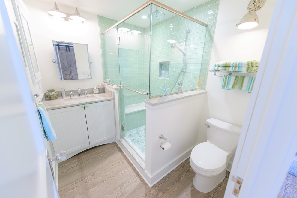This is an example of a mid-sized beach style 3/4 bathroom in Baltimore with beaded inset cabinets, white cabinets, a corner shower, a one-piece toilet, blue tile, glass tile, white walls, light hardwood floors, an undermount sink, engineered quartz benchtops, brown floor and a hinged shower door.