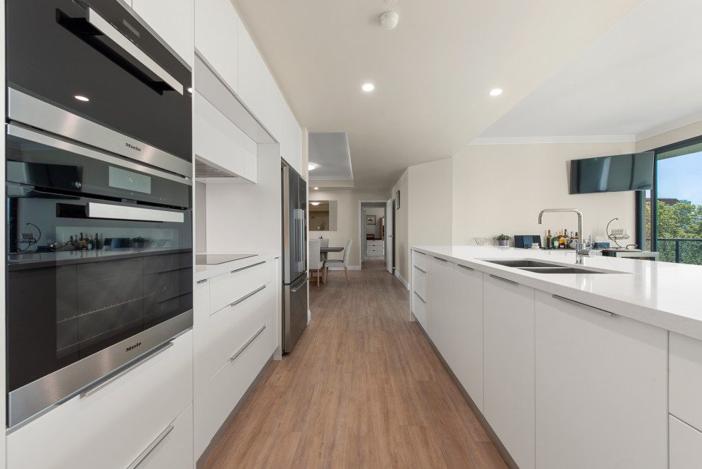 Large contemporary u-shaped open plan kitchen in Perth with an undermount sink, shaker cabinets, white cabinets, solid surface benchtops, white splashback, glass sheet splashback, stainless steel appliances, medium hardwood floors, no island and white benchtop.
