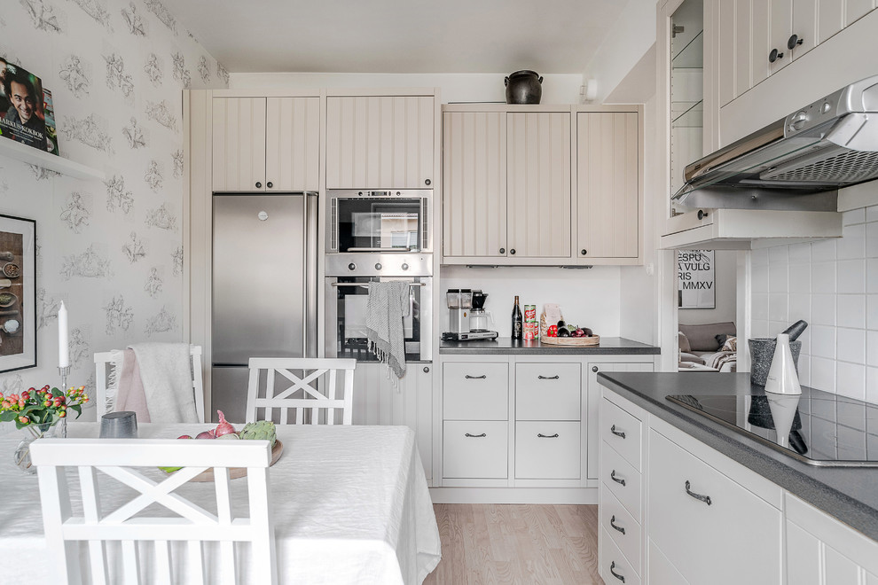 Photo of a scandinavian l-shaped eat-in kitchen in Stockholm with beaded inset cabinets, beige cabinets, white splashback, stainless steel appliances, grey benchtop, light hardwood floors, no island and beige floor.