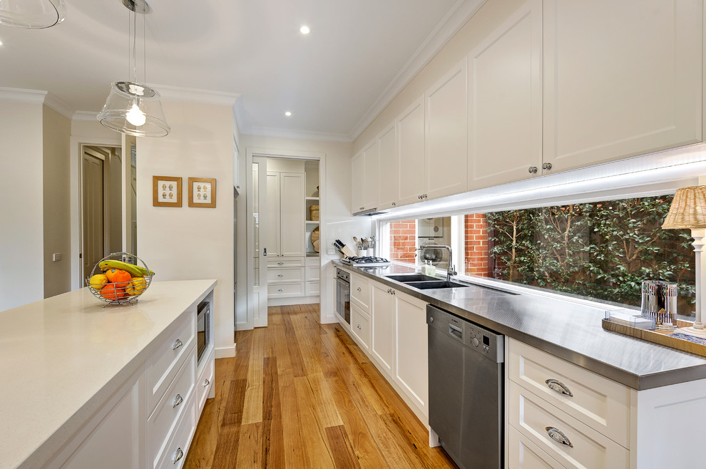 Large traditional galley kitchen in Melbourne with a double-bowl sink, white cabinets, stainless steel benchtops, stainless steel appliances, medium hardwood floors, with island and recessed-panel cabinets.