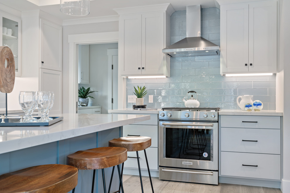 Photo of a mid-sized arts and crafts l-shaped open plan kitchen in San Francisco with a farmhouse sink, shaker cabinets, white cabinets, quartz benchtops, blue splashback, glass tile splashback, stainless steel appliances, light hardwood floors, with island, grey floor and white benchtop.