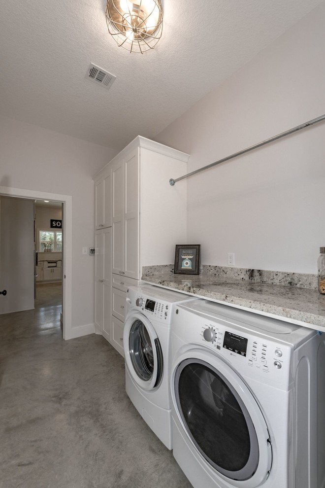 Inspiration for a mid-sized galley dedicated laundry room in Austin with a farmhouse sink, shaker cabinets, white cabinets, granite benchtops, white walls, concrete floors, a side-by-side washer and dryer and grey floor.
