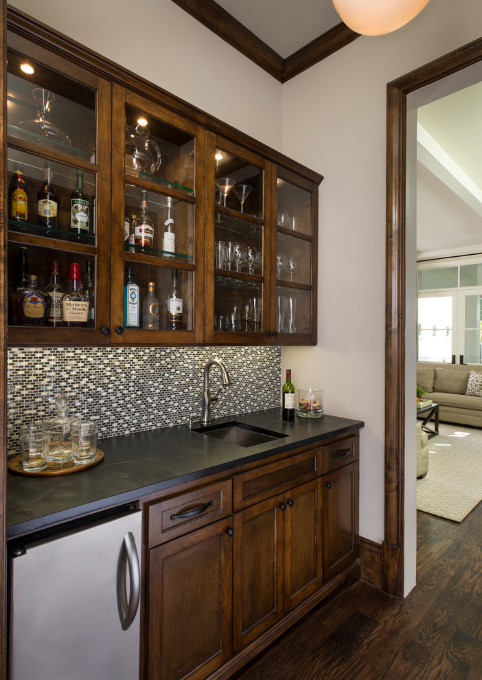 Mid-sized traditional galley wet bar in Dallas with an undermount sink, shaker cabinets, brown cabinets, soapstone benchtops, multi-coloured splashback, mosaic tile splashback and dark hardwood floors.