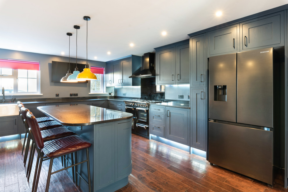 Photo of a large contemporary eat-in kitchen in Kent with a farmhouse sink, shaker cabinets, blue cabinets, quartzite benchtops, black splashback, glass sheet splashback, black appliances, dark hardwood floors, with island, brown floor and black benchtop.