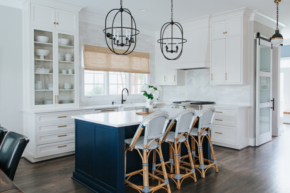 Photo of a mid-sized beach style l-shaped kitchen pantry in St Louis with a farmhouse sink, shaker cabinets, quartz benchtops, white splashback, marble splashback, white appliances, medium hardwood floors, with island, brown floor, white benchtop and blue cabinets.