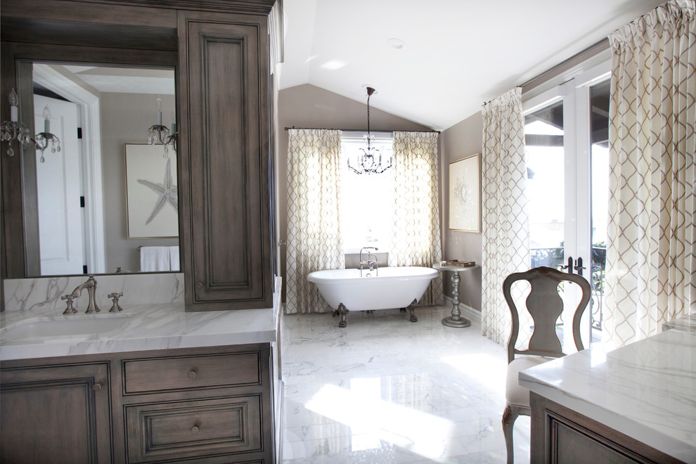 Inspiration for a large transitional master gray tile, white tile and marble tile marble floor and white floor claw-foot bathtub remodel in Orange County with recessed-panel cabinets, medium tone wood cabinets, a one-piece toilet, marble countertops, brown walls, a drop-in sink, a hinged shower door and white countertops