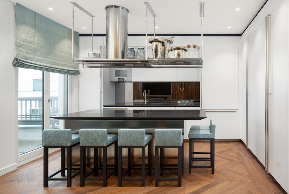 This is an example of a mid-sized contemporary l-shaped kitchen in Berlin with an undermount sink, flat-panel cabinets, white cabinets, black splashback, glass sheet splashback, panelled appliances, with island, brown floor and black benchtop.
