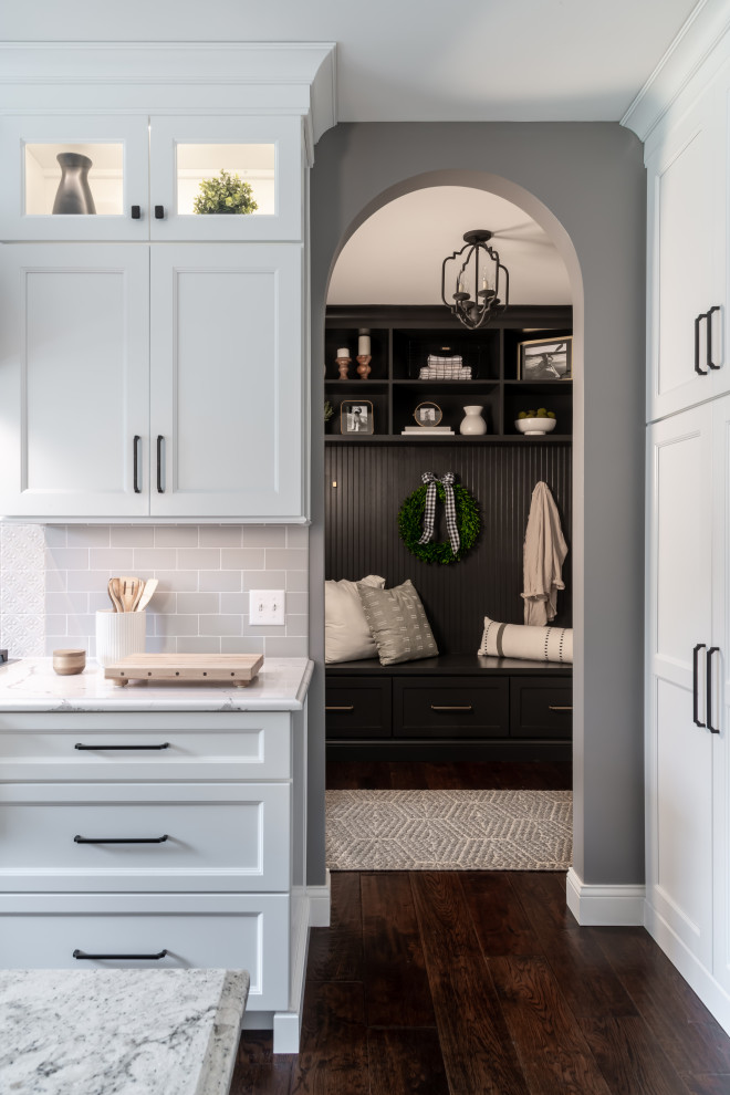 Example of a large transitional l-shaped dark wood floor and brown floor enclosed kitchen design in St Louis with a farmhouse sink, shaker cabinets, white cabinets, quartz countertops, white backsplash, subway tile backsplash, stainless steel appliances, an island and white countertops