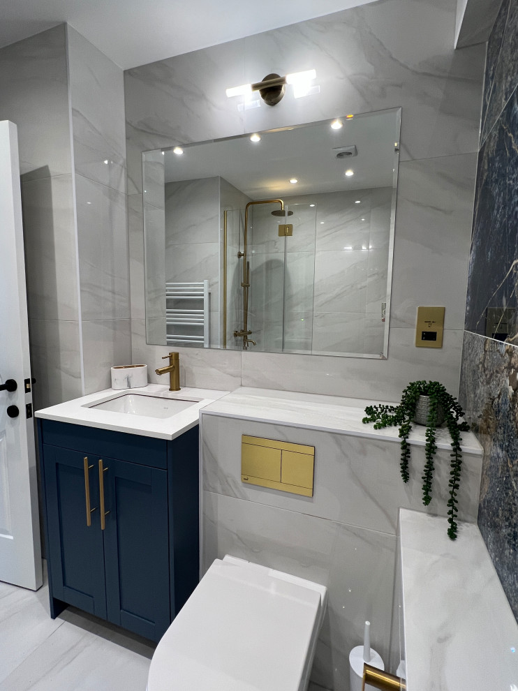 Photo of a medium sized contemporary grey and white bathroom in London with shaker cabinets, grey cabinets, a built-in bath, a shower/bath combination, a wall mounted toilet, porcelain tiles, blue walls, medium hardwood flooring, quartz worktops, brown floors, feature lighting and a single sink.