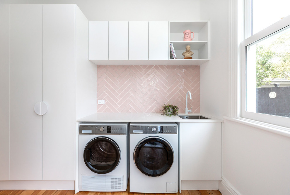 Photo of a mid-sized modern single-wall dedicated laundry room in Auckland with an undermount sink, flat-panel cabinets, white cabinets, light hardwood floors, pink splashback, subway tile splashback, white walls, a side-by-side washer and dryer and white benchtop.