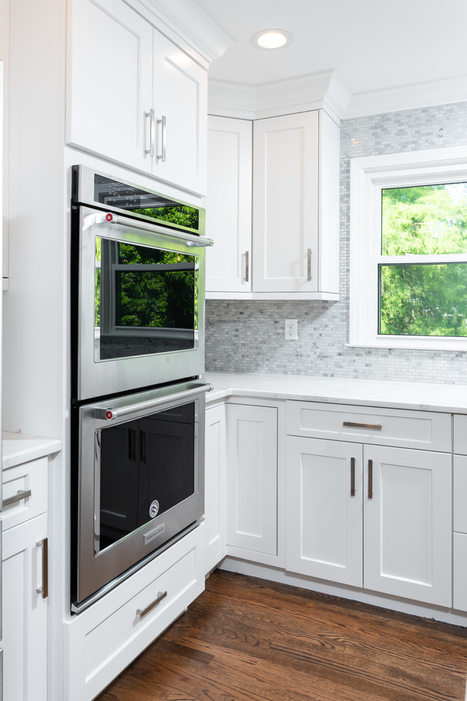 Large transitional l-shaped medium tone wood floor and brown floor eat-in kitchen photo with a double-bowl sink, shaker cabinets, white cabinets, quartz countertops, gray backsplash, mosaic tile backsplash, stainless steel appliances, an island and white countertops