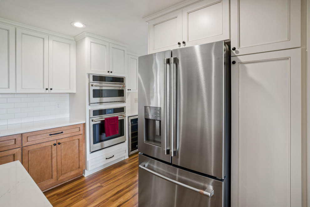 Inspiration for a mid-sized transitional l-shaped medium tone wood floor and brown floor eat-in kitchen remodel with a farmhouse sink, shaker cabinets, white cabinets, quartz countertops, white backsplash, subway tile backsplash, stainless steel appliances, an island and white countertops