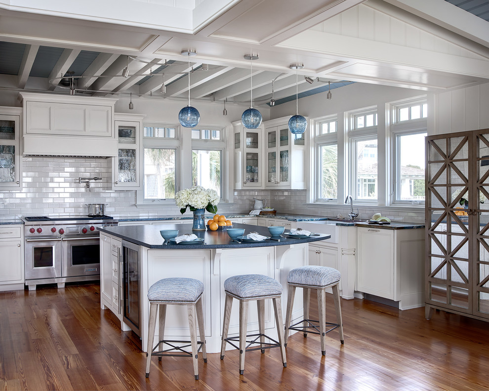 Example of a beach style l-shaped medium tone wood floor kitchen design in Raleigh with a farmhouse sink, white cabinets, white backsplash, subway tile backsplash, stainless steel appliances, an island and recessed-panel cabinets