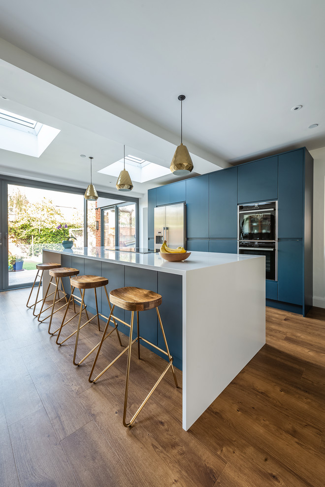 Photo of a large contemporary galley open plan kitchen in London with flat-panel cabinets, blue cabinets, quartzite benchtops, stainless steel appliances, vinyl floors, with island, white benchtop and brown floor.