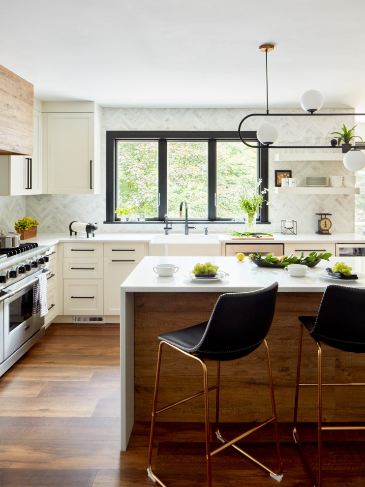 Photo of a mid-sized modern u-shaped eat-in kitchen in New York with a farmhouse sink, shaker cabinets, white cabinets, white splashback, stone tile splashback, stainless steel appliances, laminate floors, with island, brown floor and white benchtop.